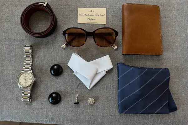 Flat lay of men's accessories: dress watch, leather belt, tie, pocket square, sunglasses and wallet