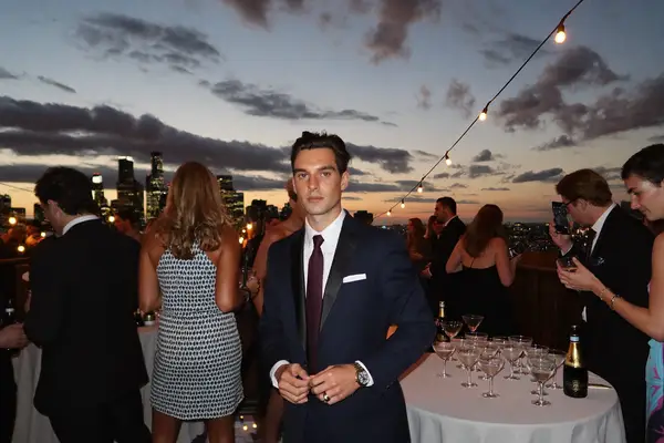 Man in a navy suit and burgundy tie at a rooftop cocktail party at dusk